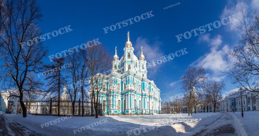 На фотографии представлен Смольный собор в Санкт-Петербурге, зимний день. Ясное голубое небо над барочным фасадом собора, украшенным многочисленными лепнинами и золотыми куполами, создаёт контраст с белым снегом, покрывающим землю и деревья без листьев. Этот снимок может быть использован в исторических и культурных изданиях, в туристических путеводителях, а также в образовательных материалах, посвященных русской архитектуре.